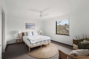 A bedroom with a bed, a ceiling fan, a large window, a woven rug, and a wicker chair.