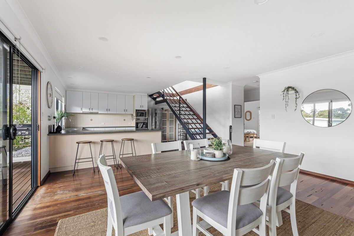 A modern kitchen with white cabinets, a wooden dining table, and a staircase leading to an upper level.