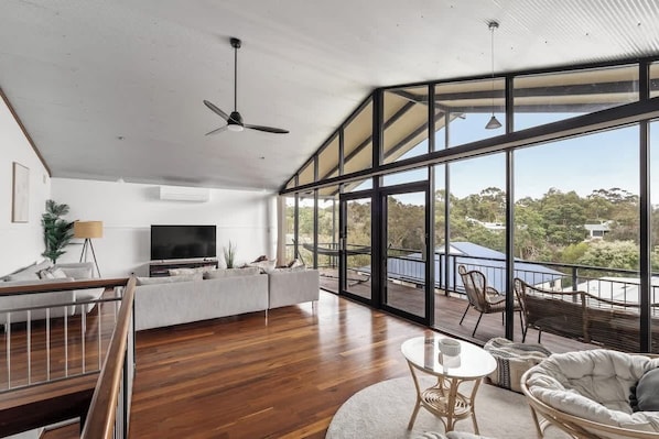 A spacious living area with a large TV, wooden flooring, a balcony with outdoor seating, and a ceiling fan.