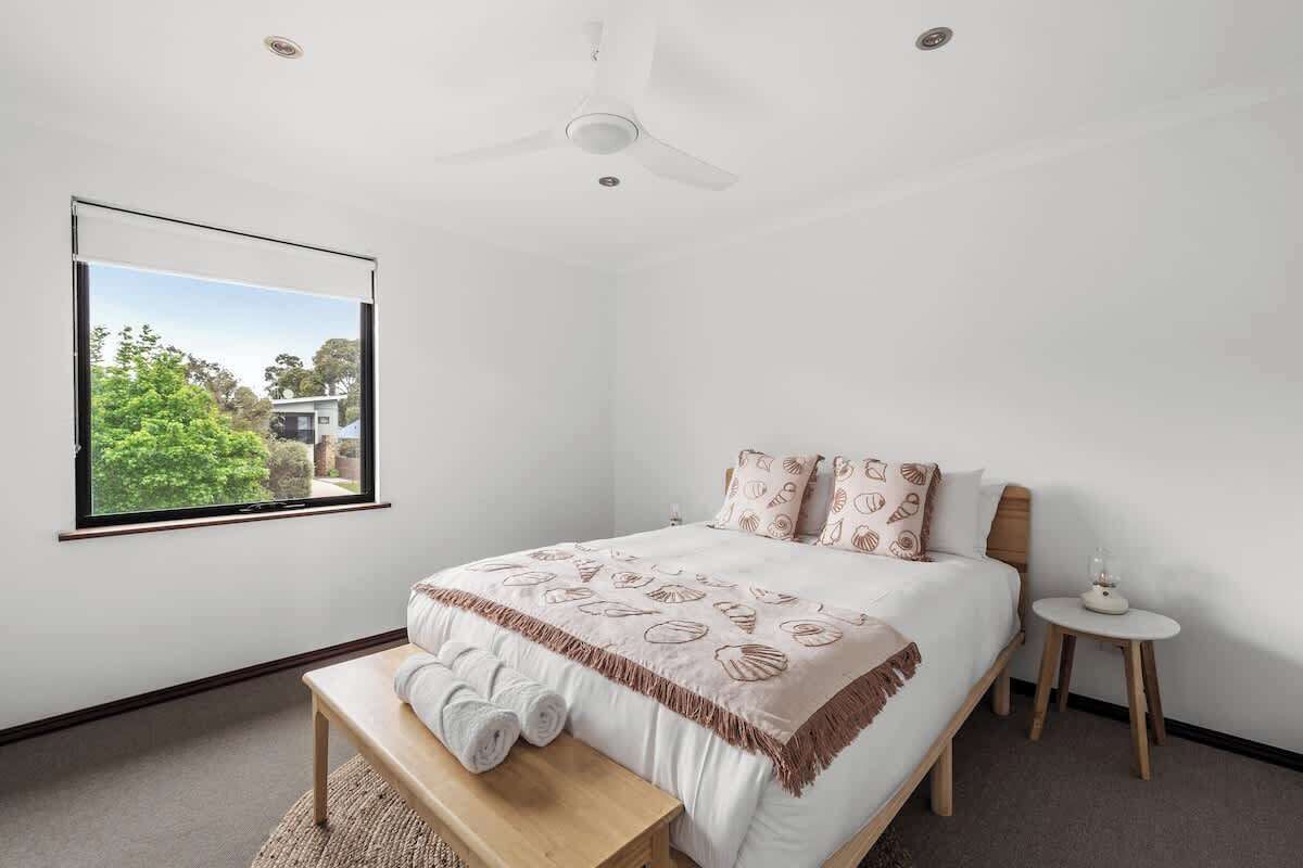 A bedroom with a bed, a bench, a small table, a ceiling fan, and a window with a view of greenery.