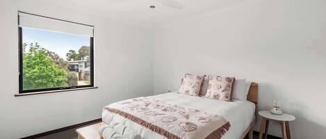 A bedroom with a bed, a bench, a small table, a ceiling fan, and a window with a view of greenery.