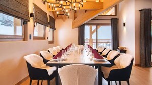 Un comedor con una mesa larga dispuesta para una comida, rodeada de sillas con mantas de piel, un candelabro y ventanas amplias con cortinas.