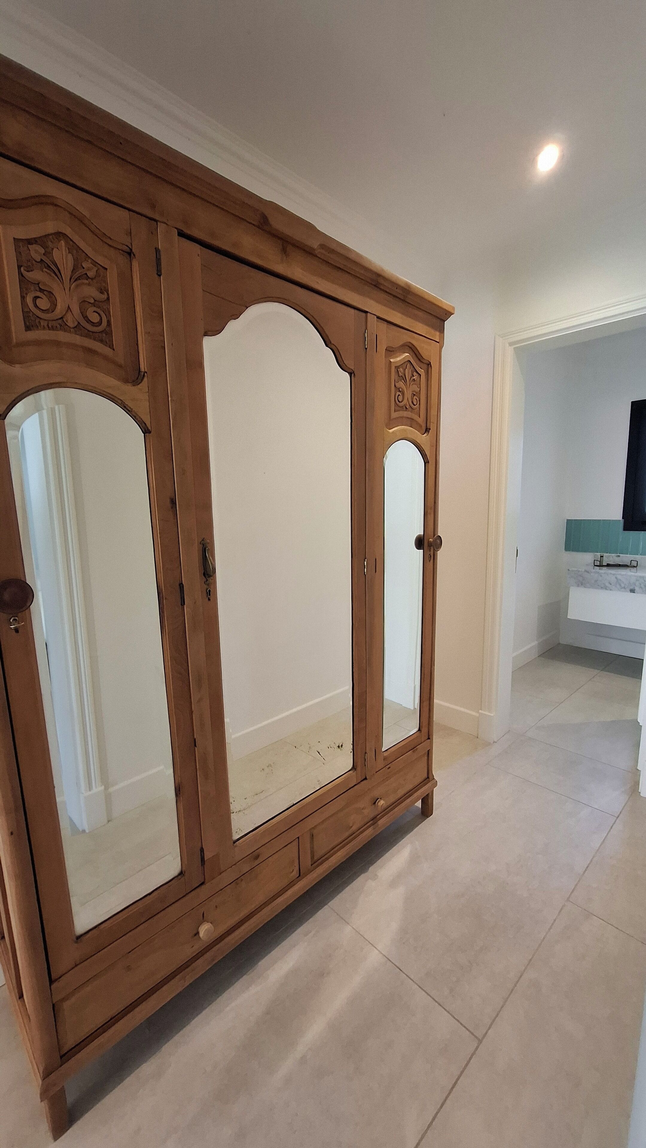 Guarda-roupa de madeira com portas espelhadas e uma gaveta pequena, situado num corredor com soalho em azulejos e parede branca.