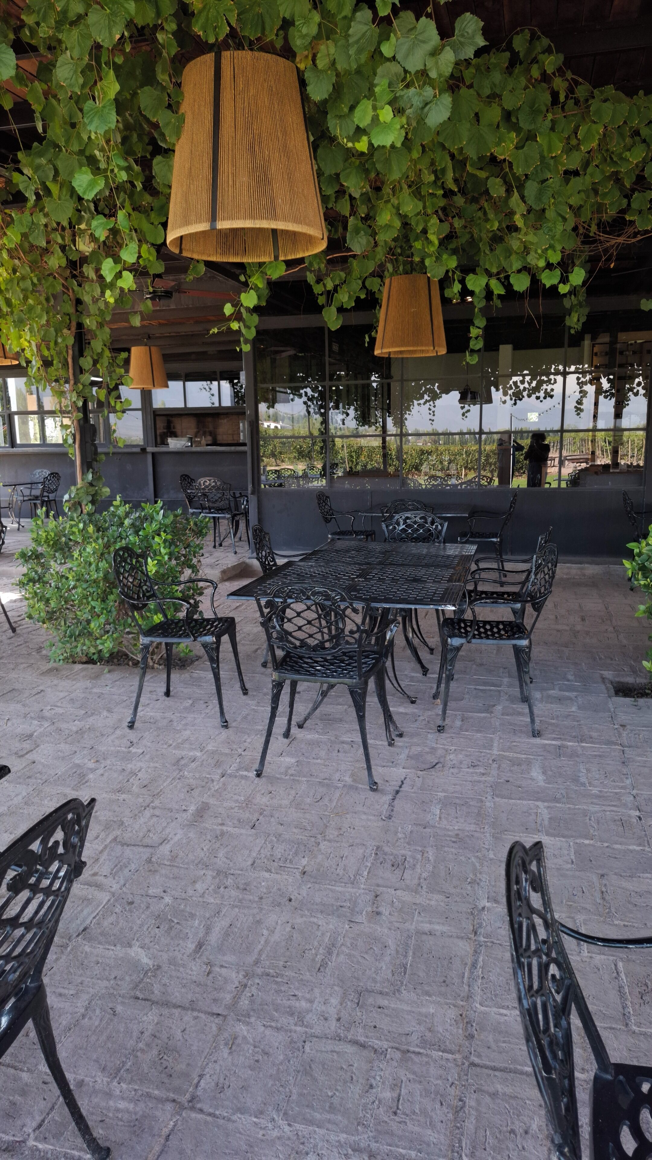 Un patio al aire libre con una mesa y sillas de metal, rodeado de follaje y lámparas colgantes.