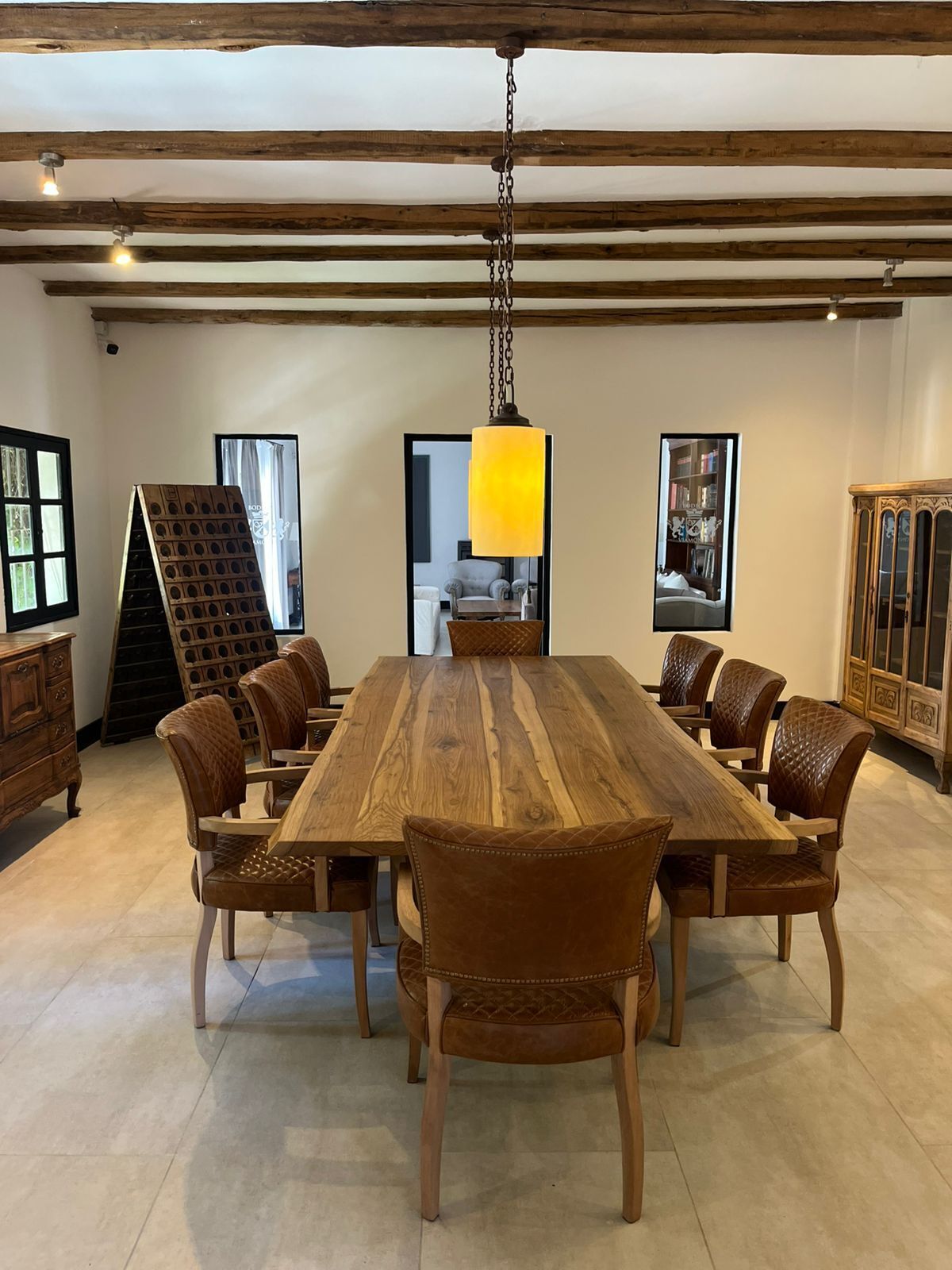 A dining room with a wooden table and leather chairs, a hanging light fixture, and a view of a living area through a window.