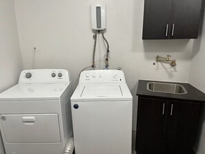 A laundry area with two white Whirlpool washers, a black cabinet with a sink, and a wall-mounted water heater. - 3-bedroom house with Solar Energy, Water cistern, AC, and high speed internet. (Canaboncito)