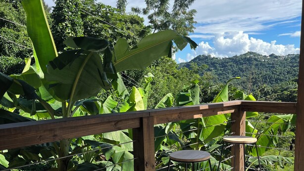 A wooden deck with two metal bar stools and a wooden table, surrounded by lush greenery and a view of a hilly landscape. - 3-bedroom house with Solar Energy, Water cistern, AC, and high speed internet. (Canaboncito)