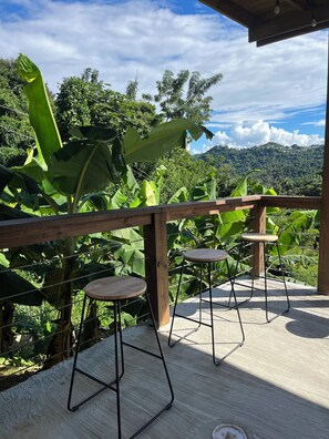 A wooden deck with two metal bar stools and a wooden table, surrounded by lush greenery and a view of a hilly landscape. - 3-bedroom house with Solar Energy, Water cistern, AC, and high speed internet. (Canaboncito)