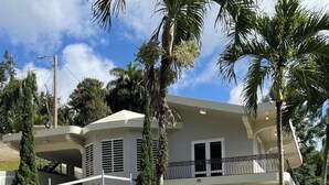 A two-story house with a balcony, surrounded by palm trees and a well-maintained lawn. - 3-bedroom house with Solar Energy, Water cistern, AC, and high speed internet. (Canaboncito)