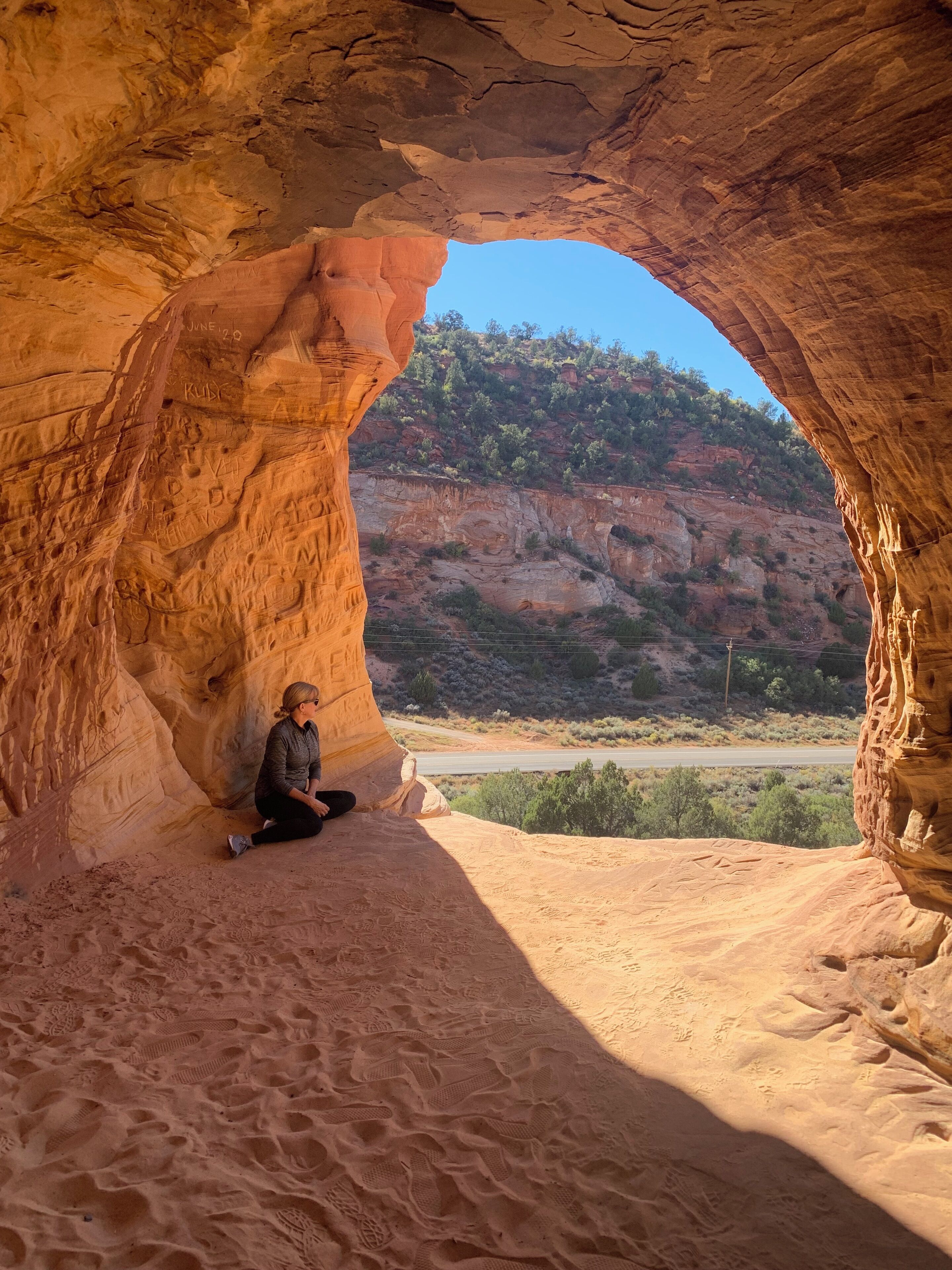 Une personne est assise à l’intérieur d’une arche naturelle formée par des roches, avec un paysage désertique et une route en arrière-plan.