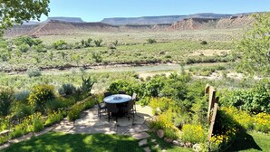 Eine Terrasse mit rundem Tisch und Stühlen, umgeben von Grünpflanzen und Blick auf einen Fluss und Berge.