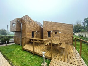 A wooden cabin with a deck and a small attached structure, featuring a door and windows. - Pretty 12-person gîte in Fiefs (Ternois 62) 3 épis, ideal for hiking/biking (Fiefs)