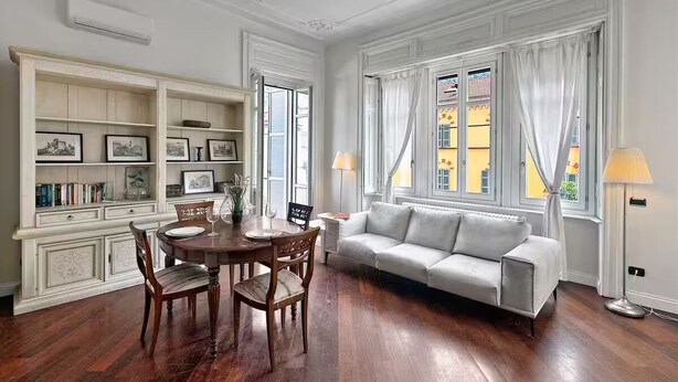 A dining area with a wooden table and chairs, a white sofa, and a bookshelf with framed pictures. - Historic Villa in Como Center + 4 free parking (Como)