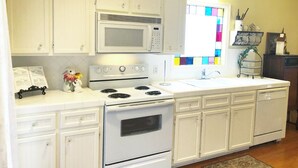 Cocina con gabinetes blancos, estufa blanca y microondas. Hay una ventana con vidrio vidriado colorido, y un estante pequeño con objetos decorativos.