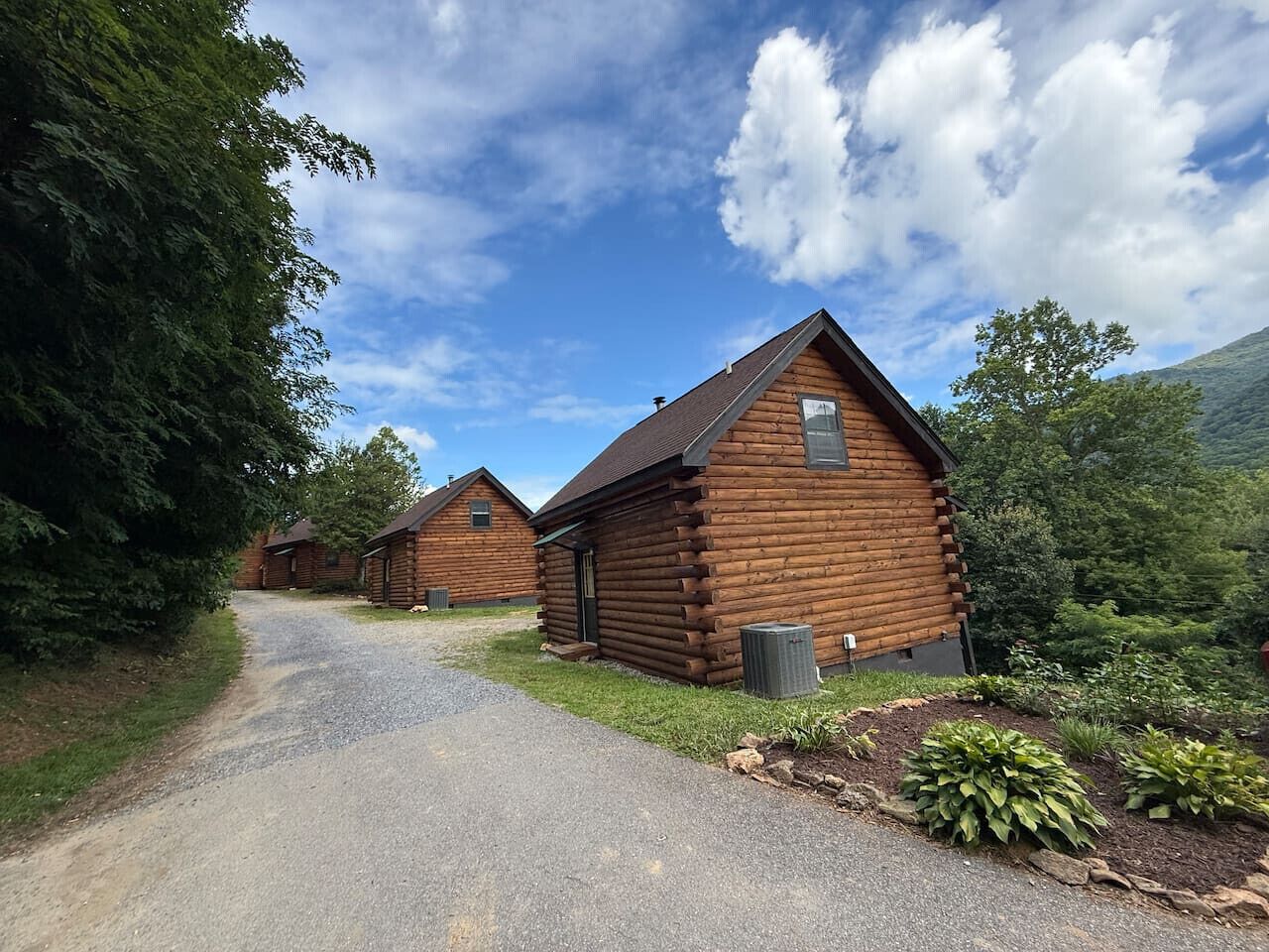 A row of log cabins with a gravel driveway leading to them, surrounded by greenery and hills.