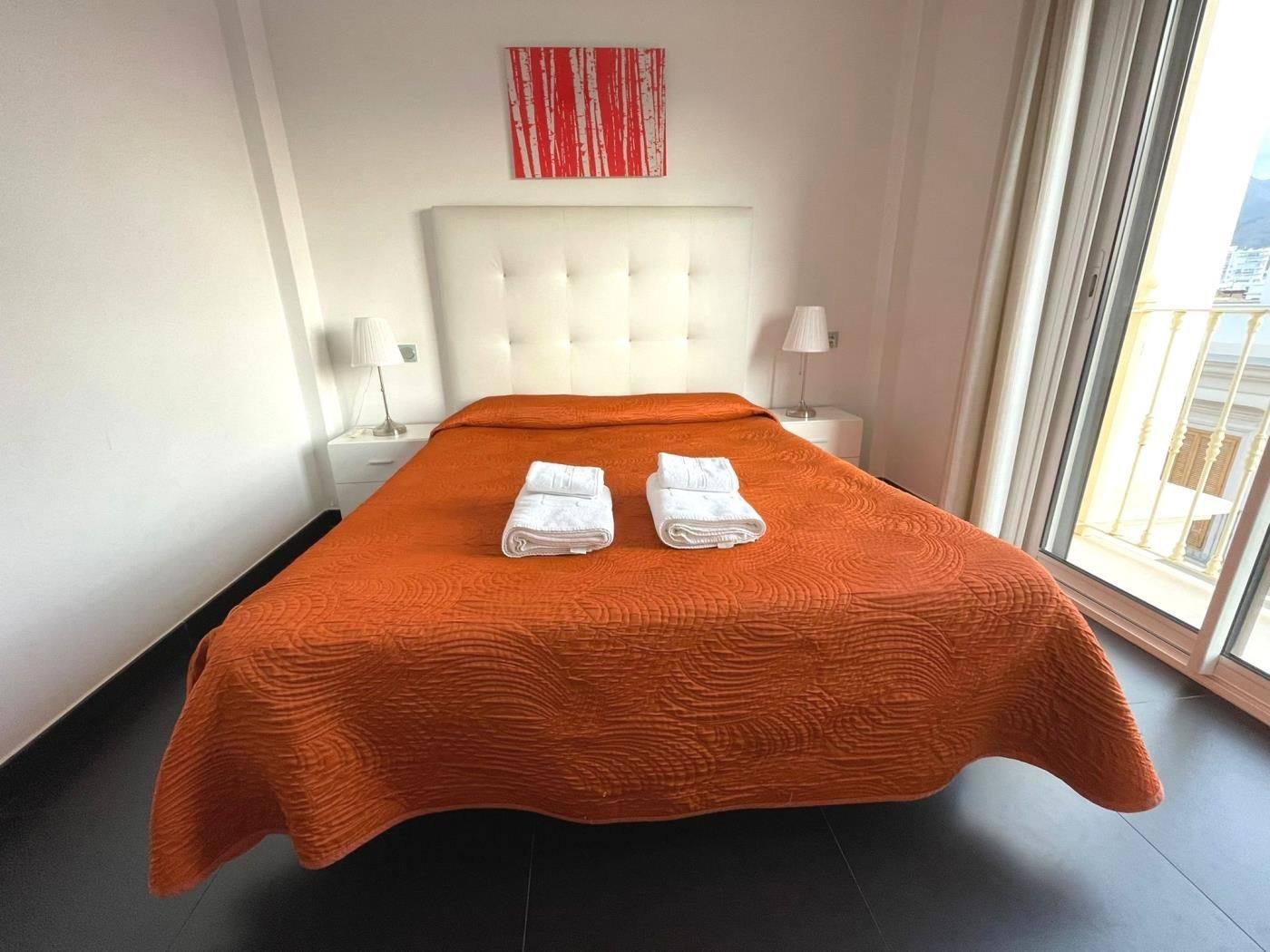 A hotel room with a large bed, two white towels, a white headboard, a window with a view, and a red abstract painting on the wall.