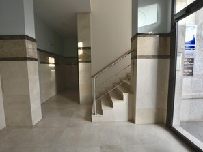 Un pasillo con piso de mármol, una escalera metálica y una puerta de cristal con señalización.