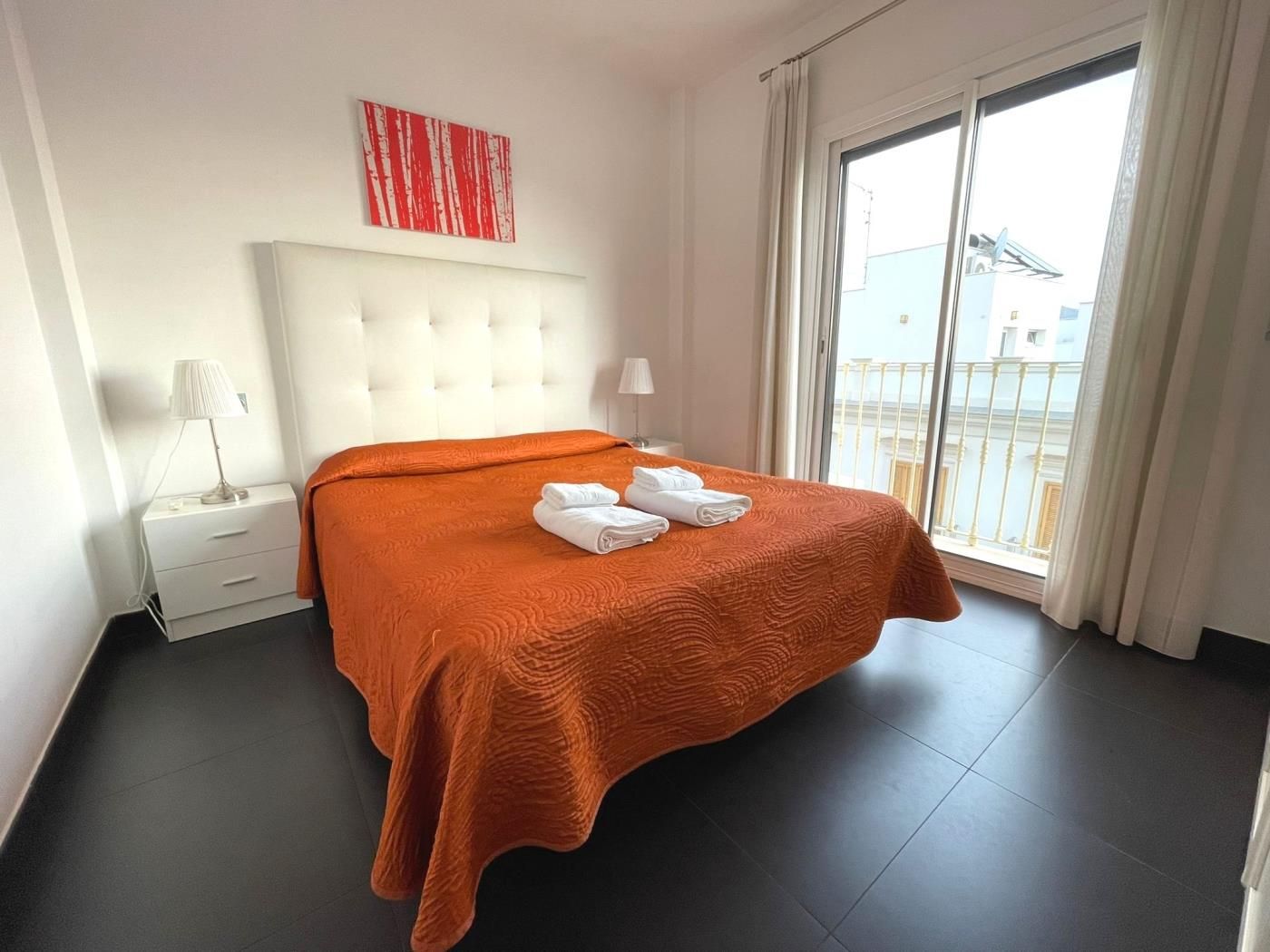 A hotel room with a large bed, a white headboard, a window with a view of buildings, and a red abstract painting on the wall.