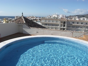 A swimming pool with clear blue water, surrounded by a white building with a tiled roof, and a view of a coastal cityscape in the background. - Modern one bedroom apartment in the heart of Nerja (Nerja)
