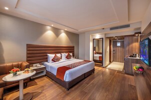 A modern hotel room with a large bed, a seating area, a television, and a small kitchenette.