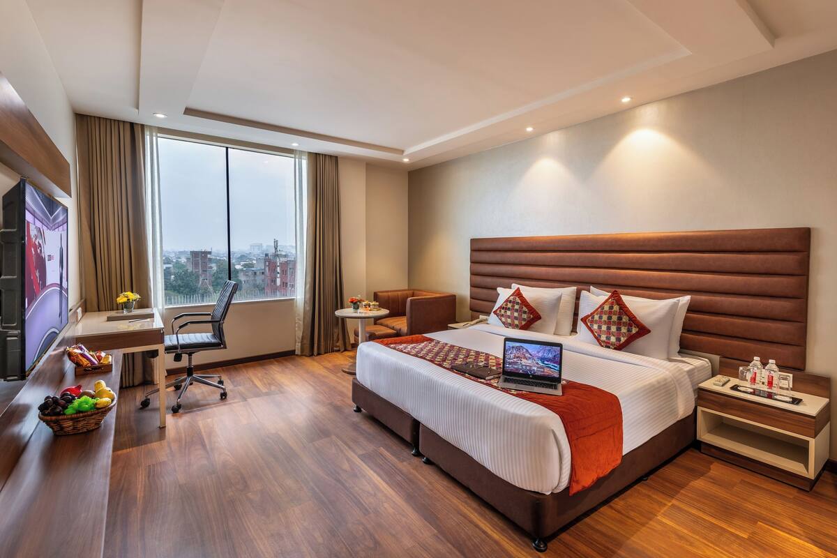 A modern hotel room with a large bed, a desk with a laptop, a TV, and a city view.