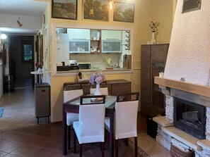 Comedor con una mesa preparada para cuatro personas, chimenea y cocina visible a través de una ventana.