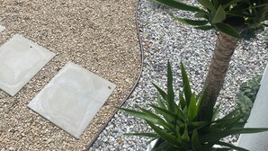 Ein Gartenbereich mit Kiesgrund, einer Kakteepflanze und einem Blumentopf mit langen, grünen Blättern.