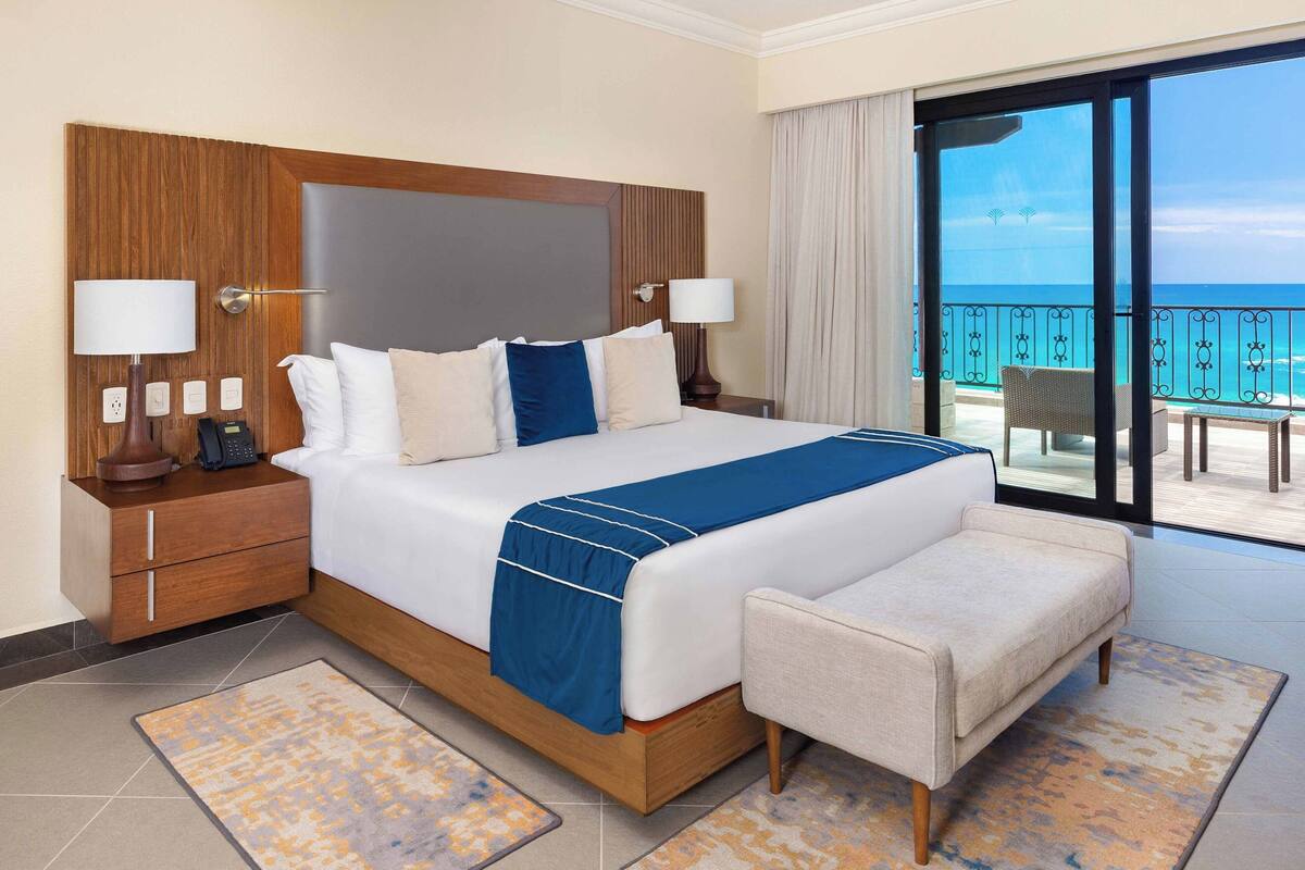 Ein Hotelzimmer mit einem großen Bett, einem Nachttisch, einem Schreibtisch, einem Sessel und einem Balkon mit Meerblick.