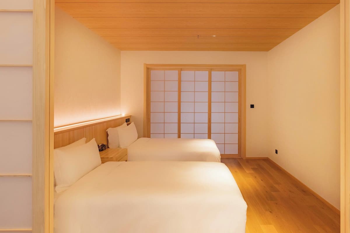 Quarto com duas camas, piso de madeira, porta deslizante e mesa de cabeceira com abajur.