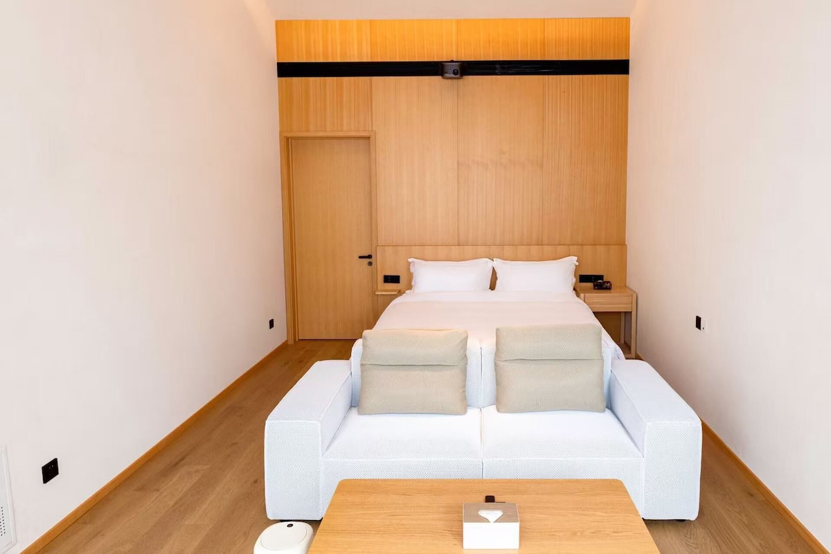 Quarto de hotel pequeno com uma cama de solteiro, uma mesinha de cabeceira e um sofá.
