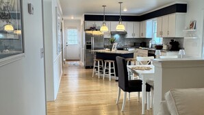 A modern kitchen with white cabinets, a central island with stools, pendant lighting, and a dining area with a white table and chairs. - The Retreat at Bethany Beach Cove (Sussex County)