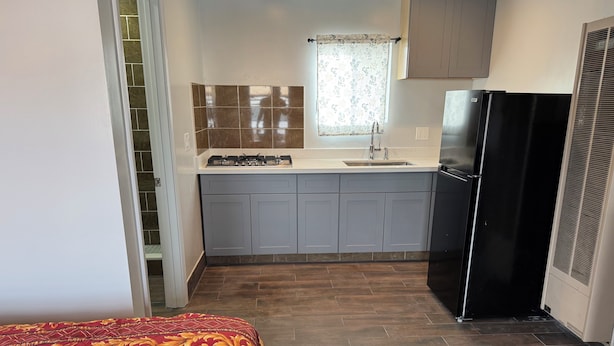 A compact kitchen with grey cabinets, a black refrigerator, and a window with floral curtains. - BORONDA INN (Salinas)