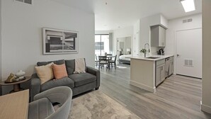 A modern apartment with a grey sofa, a dining area, and a kitchen with white cabinets. - Landing | Modern 2BD, Clubhouse, Game Room (Tempe)