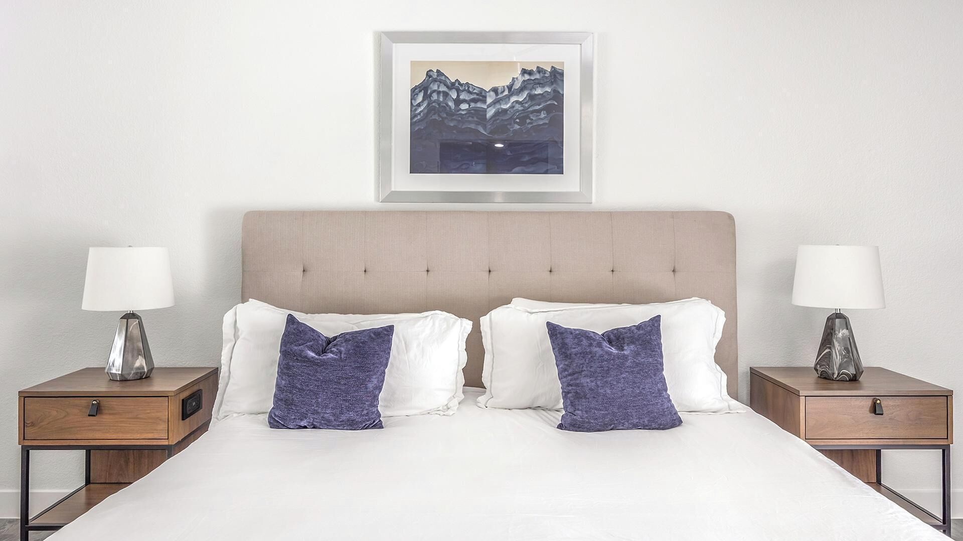 Un lit double avec une tête de lit capitonnée, deux tables de chevet avec des lampes et un tableau encadré représentant des montagnes accroché au mur.