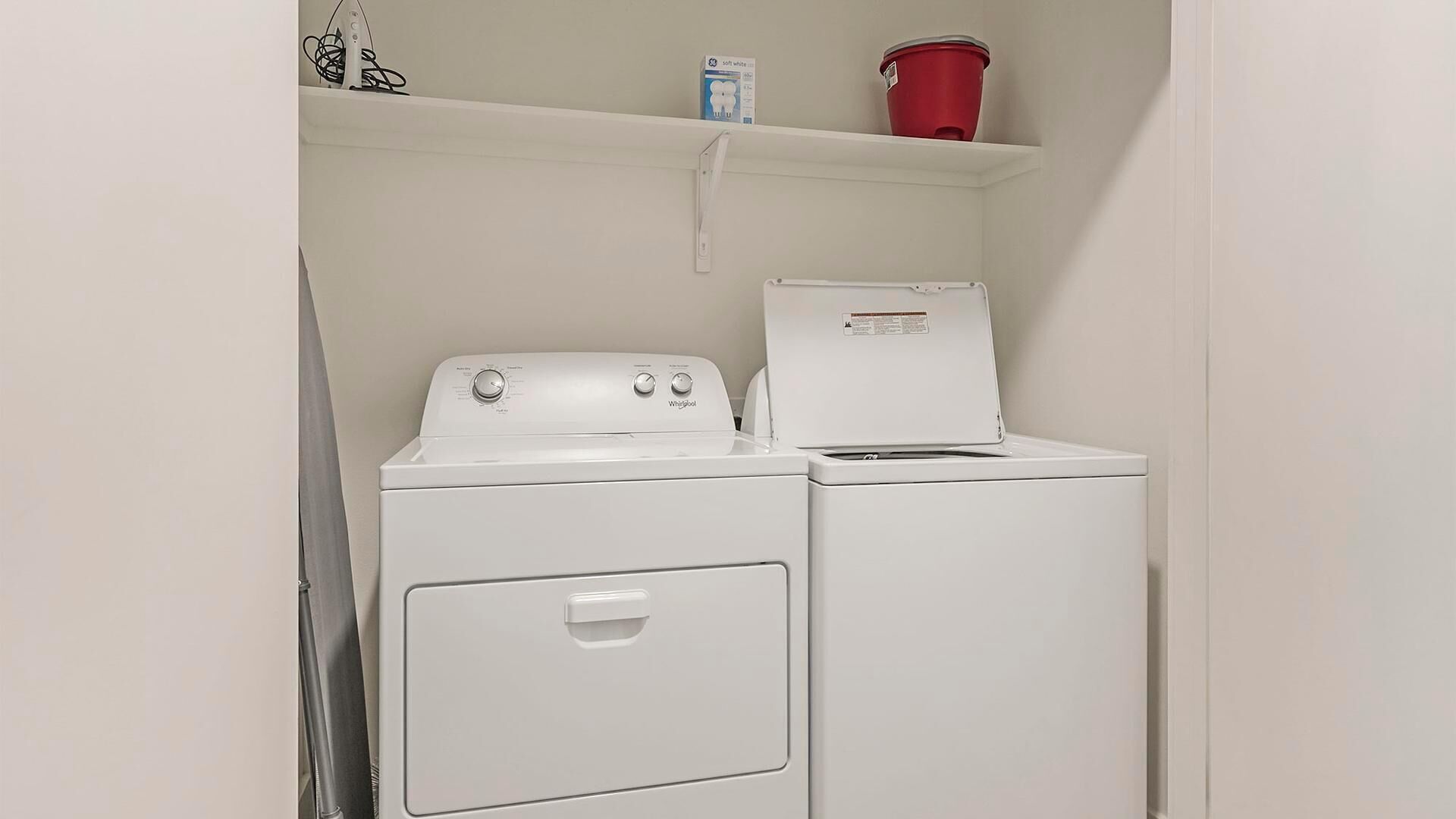 Een kleine wasruimte met een voorlader wasmachine en droger, een plank met een rode emmer en een doos wasmiddel.