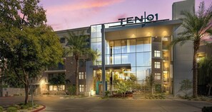 Ein modernes Hotel mit Glasfassade, mit der Aufschrift "TEN01", einem gut beleuchteten Eingang und umliegender Bepflanzung.