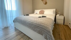 Un dormitorio con una cama grande, una mesita de noche, una decoración en la pared y una ventana con cortinas de gasa.