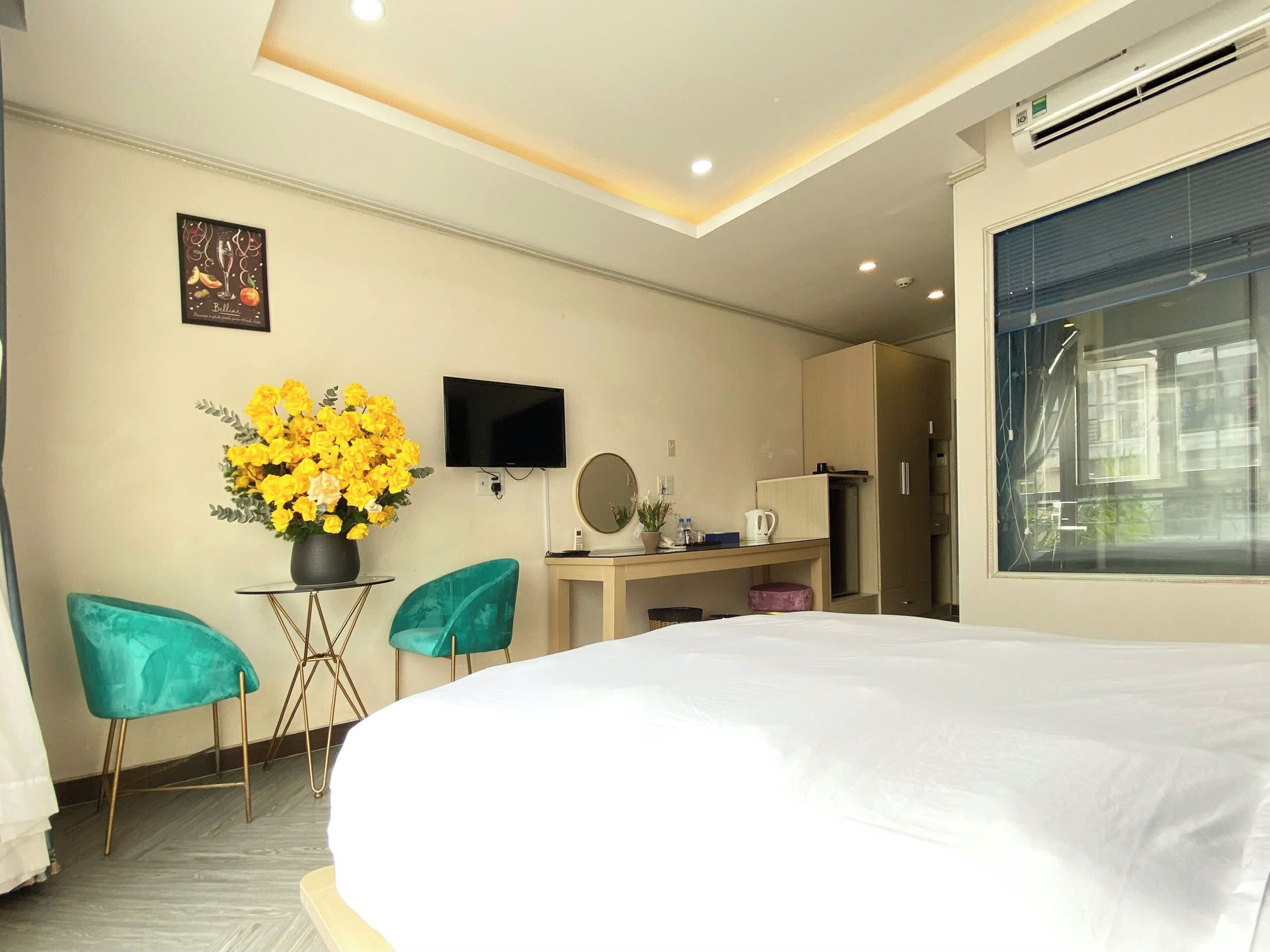 A hotel room with a large bed, a desk with a flat-screen TV, a small table with a vase of flowers, two teal chairs, and a large window with a view of buildings.