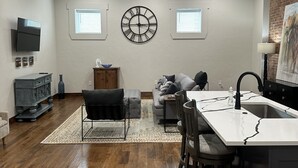 Una cocina moderna con una isla central, una zona de comedor con mesa y sillas, un sofá gris y un reloj de pared grande.