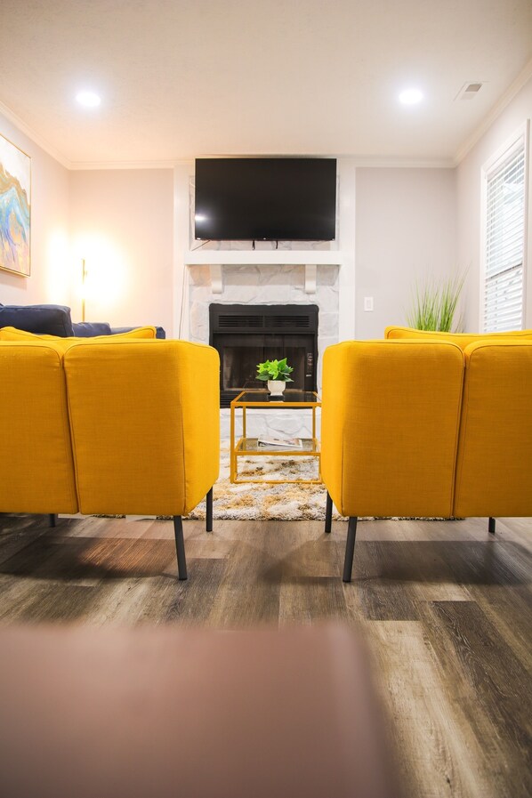 A living room with a fireplace, two yellow sofas, a flat-screen TV, and a small table with a plant. - Fayetteville Elite Escape – Close to Base & VA Hospitals (Fayetteville)