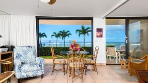 A modern room with a dining area, a large window with a view of the ocean and palm trees, and a ceiling fan. - Zen by the Sea Ground-level Paradise With Ocean Views Whales Watching (Lahaina)