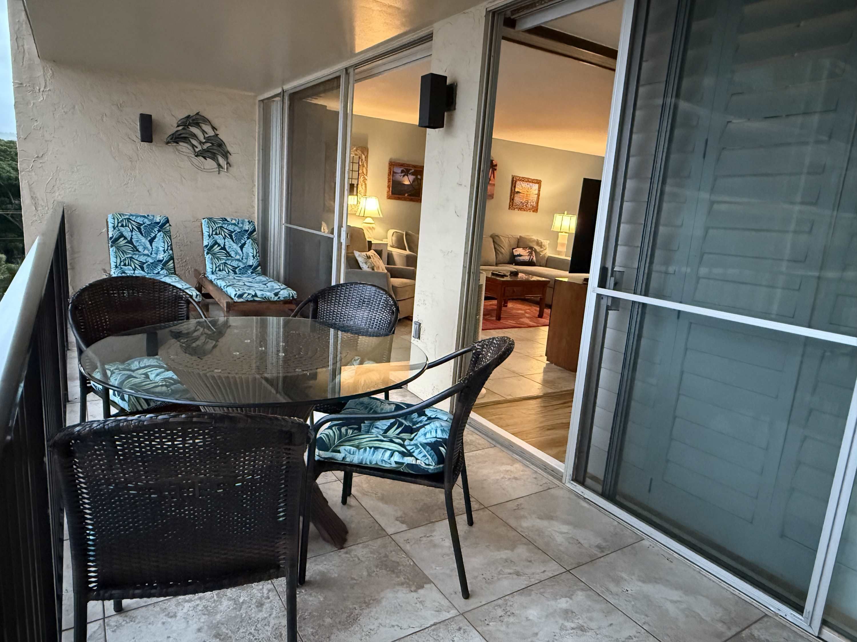 Un balcon avec une table et des chaises en verre, une porte-fenêtre coulissante donnant sur un espace de vie intérieur comprenant un canapé et une lampe.