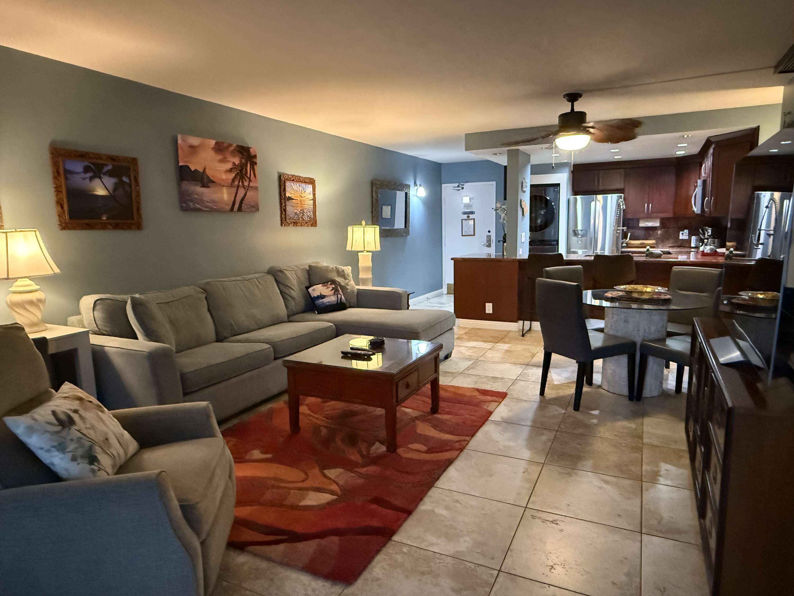 Un salon comprenant un canapé, une table basse et un coin repas avec des chaises et une table. On y trouve également un coin cuisine avec des placards et un réfrigérateur à l’arrière-plan.