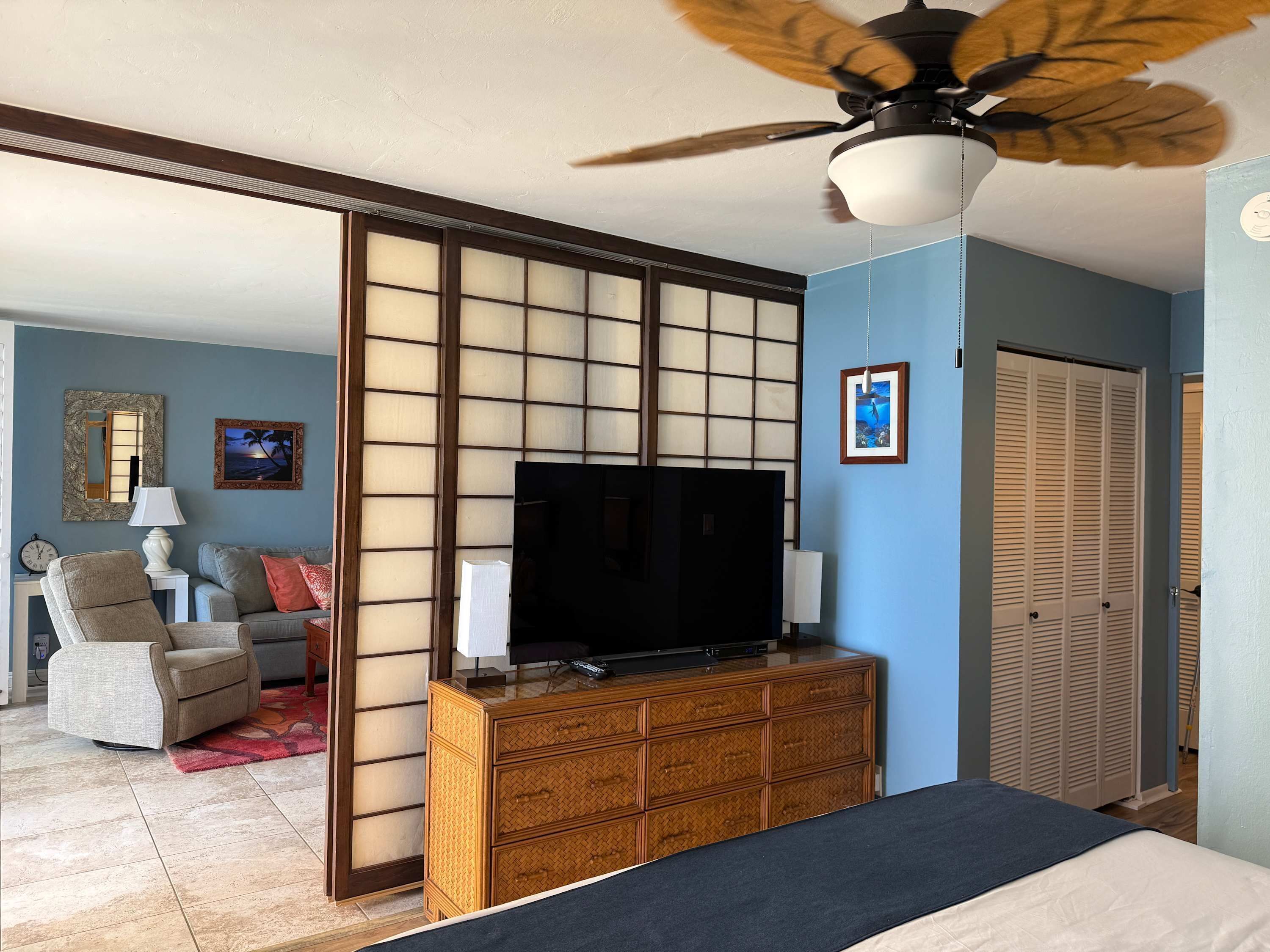 Habitación de hotel con cama, televisor sobre una cómoda de madera, ventilador de techo y un fusuma corredero.