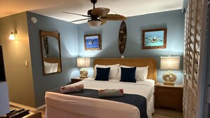 A hotel room with a bed, two bedside tables, a ceiling fan, and wall-mounted artwork. - Whale Watching Oceanfront Escape Molokai Views Cool Tropical Breezes (Lahaina)