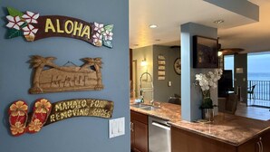 A kitchen with a wooden island, a view of the ocean, and decorative wall art including a sign that says "ALOHA" and a wooden sign with a palm tree. - Whale Watching Oceanfront Escape Molokai Views Cool Tropical Breezes (Lahaina)