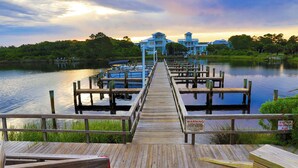A wooden pier leading to a dock with boats, surrounded by lush greenery and a large building in the background.