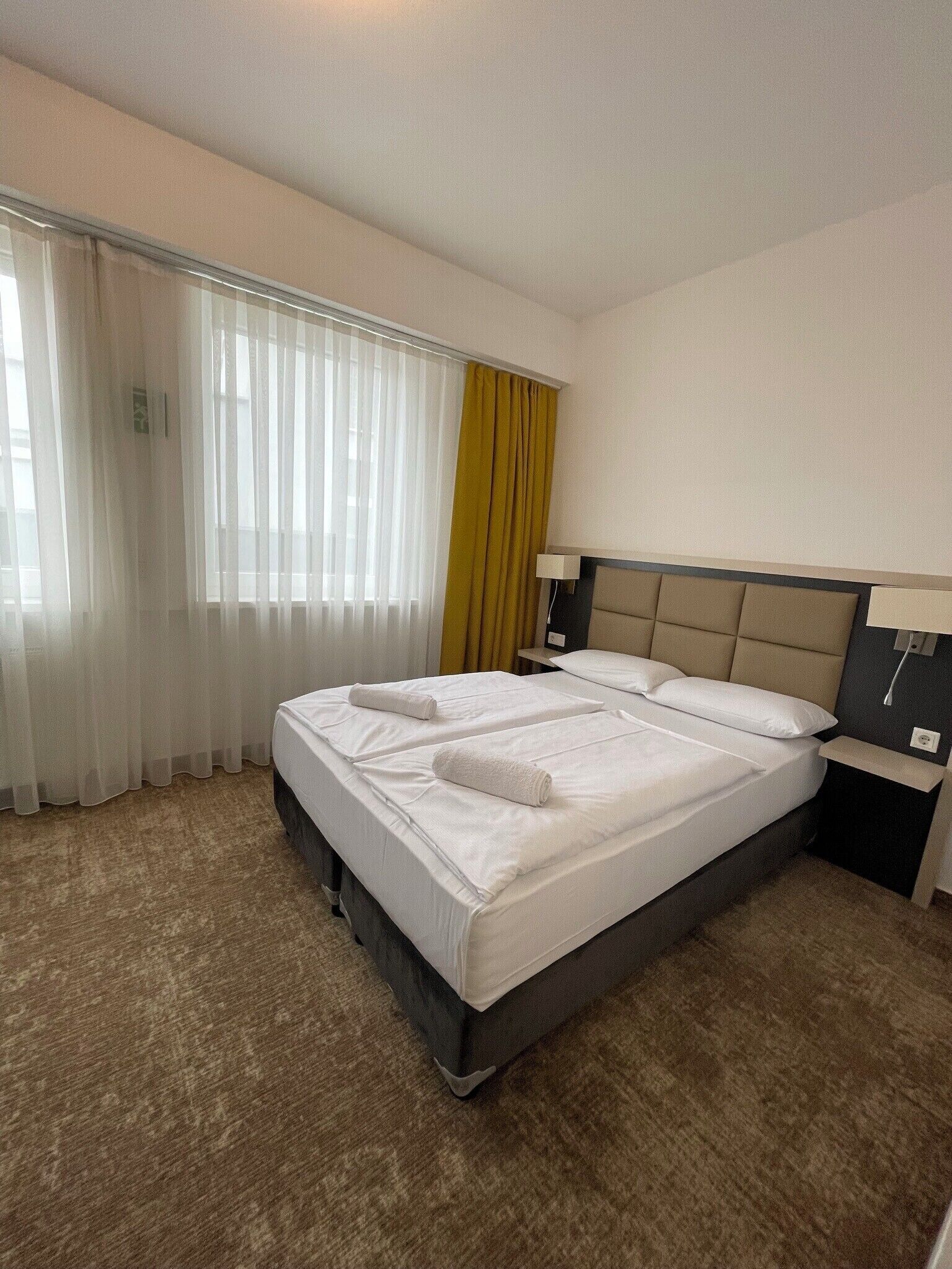 A hotel room with a large bed, two bedside tables, a headboard, and a window with sheer curtains and a yellow drape.