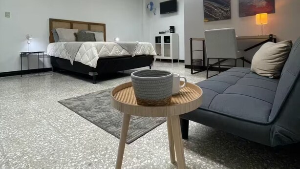 A modern bedroom with a bed, a grey sofa, a small round table with a cup, and a TV on the wall. - Quaint Downtown Loft/WI-FI (San José)
