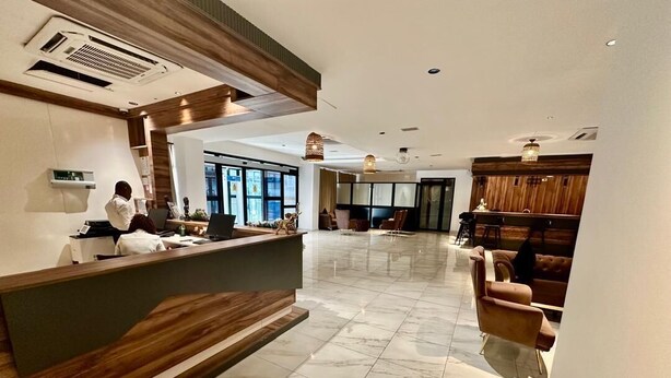 A hotel reception area with a wooden reception desk, a waiting area with leather chairs, and a large window providing a view of the outside. - Hôtel La Pivoine (Abidjan)